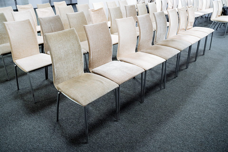 In einem geräumigen Raum sind Stühle ordentlich in Reihen aufgestellt und schaffen eine einladende Atmosphäre für Versammlungen. Die Stühle stehen auf dunkelgrauem Teppichboden.